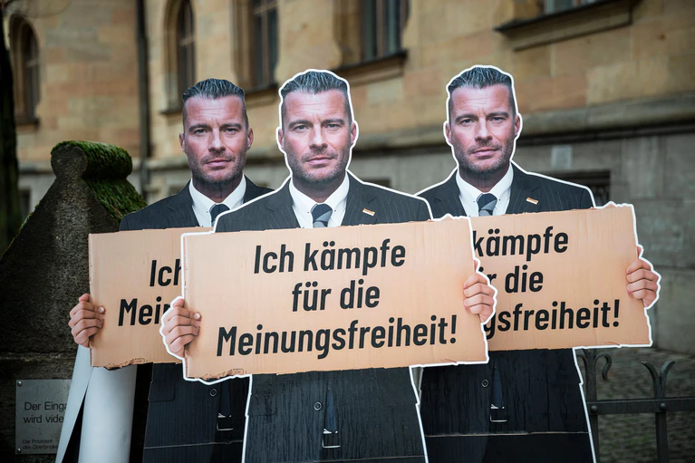 Vor dem Landgericht Bamberg stehen Pappaufsteller, die den Journalisten David Bendels mit einem Schild in der Hand zeigen - er selbst bezeichnete seinen Post als Satire. — © Daniel Vogl/dpa