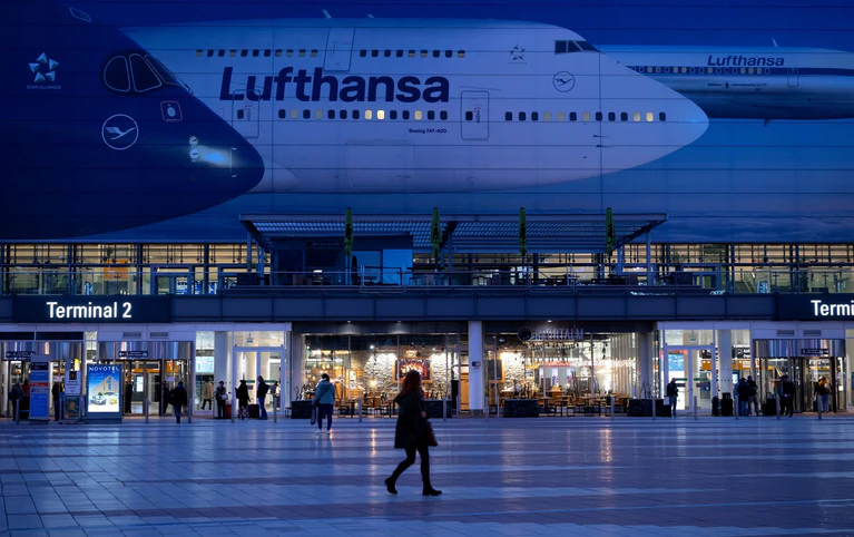 Passagiere gehen am Münchner Flughafen vor dem Terminal 2 an einem Plakat mit Flugzeugen der Lufthansa entlang. — © Sven Hoppe/dpa