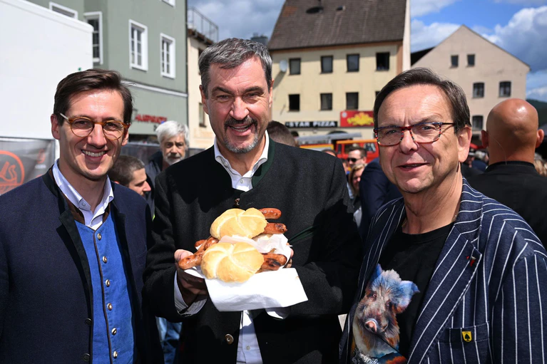 Markus Söder (M.) hält Bratwurst-Semmeln in die Kamera. — © Felix Hörhager/dpa