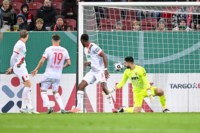 Der Schuss, der zum Augsburger Pokal-K.o. führt. Verteidiger Chrislain Matsima (M) fälscht den Ball auch noch unglücklich und damit unhaltbar ab.  — © Harry Langer/dpa